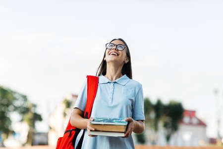 A cheerful student girl with a backpack and books, standing and smiling on the streetの写真素材
