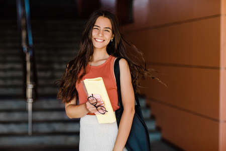 Portrait of a charming student girl with a smile, holding a folder, glasses and a phoneの写真素材