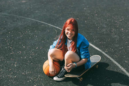 A smiling girl with a basketball is sitting on a skateboard on the sports fieldの写真素材