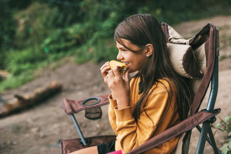 A teenage girl in a bright yellow sweater is sitting in a camping chair on the shore of the lake and eating cornの写真素材