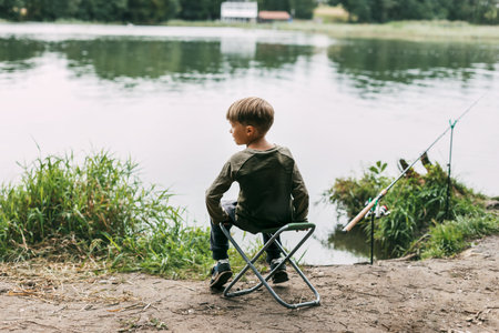 The boy is sitting on a folding chair on the shore of a lake or river. Recreation, weekends, tourism. Rear viewの写真素材