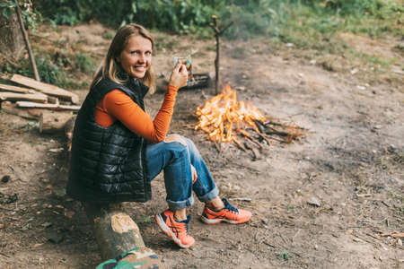A young woman is warming herself by the fire with a cup of warming tea in the forest