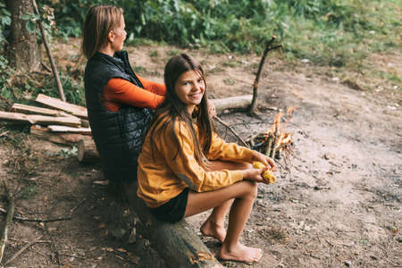 A mother with her teenage daughter is sitting on a tree near a campfire in the forest or park during...
