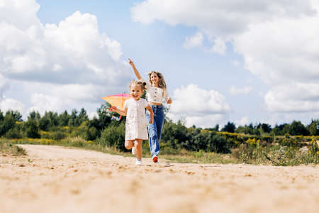 A laughing little girl and her mother run along the road to the field and fly a kite. Mom and daughter have fun togetherの写真素材