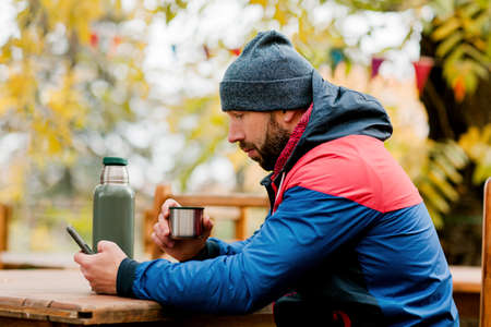 A young man using a mobile phone is sitting outdoors in a cafe or restaurant on an autumn day,...