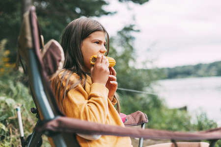 A teenage girl in a bright yellow sweater is sitting in a camping chair on the shore of the lake and eating cornの写真素材