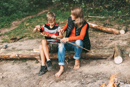 A mother and her teenage son are sitting on a tree in campsites and trying grilled sausages cooked on a fireの写真素材