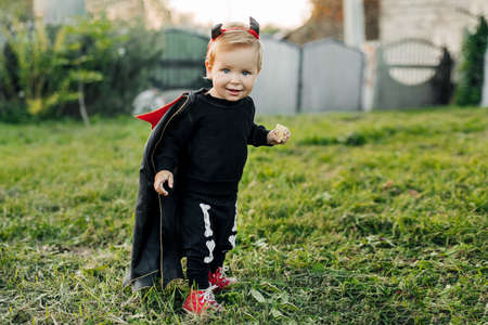 A charming kid in a devil costume and a raincoat sneaks around the garden. Halloween concept, carnival costumeの写真素材