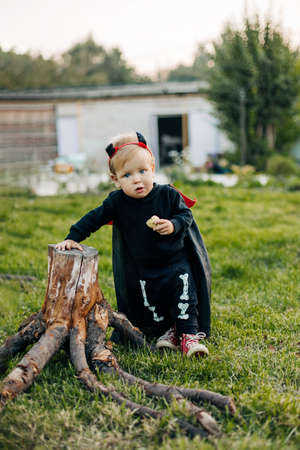 A blond boy in a devil costume with a red cloak stands near a tree stump. Halloween eve, carnival costume, traditionsの写真素材