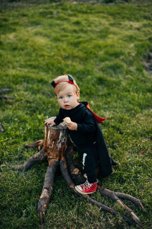 A blond boy in a devil costume with a red cloak stands near a tree stump. Halloween eve, carnival costume, traditionsの写真素材