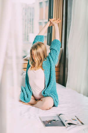 A young girl in pyjamas and a blue knitted cardigan is sitting cross-legged on the bed, stretching. The girl enjoys the morning and enjoys the new dayの写真素材
