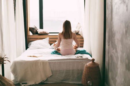 A young woman in pyjamas is sitting on a bed in a Lotus position with her fingers folded in a mudra gesture, enjoying a morning of deep mindfulness meditation. Rear viewの写真素材