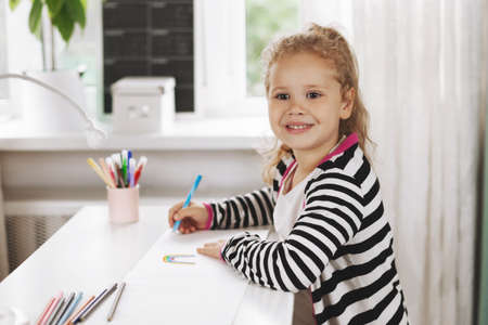 Portrait of a charming girl who draws a rainbow in an album with colored pencils. Homework, home training, social distance. Side view, space for textの写真素材
