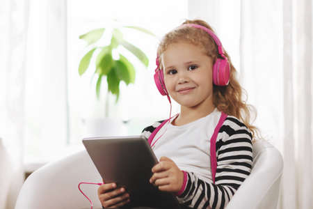 A school girl in pink headphones sits with a tablet and listens to an online lesson. Online education, online training, social distanceの写真素材