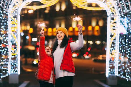 A beautiful boy and his mother celebrate New Year or Christmas with sparklers on the background of a night city with illuminated illuminationの写真素材