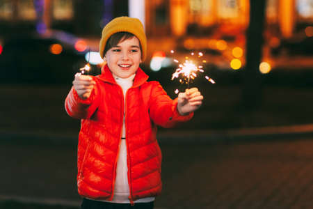 A charming boy holds a sparkler in his hands, celebrating the new year at night on the street. The...