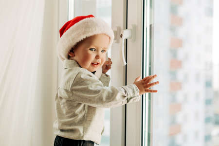 A cute little boy in a Santa hat is standing at the window in his house and smilingの写真素材