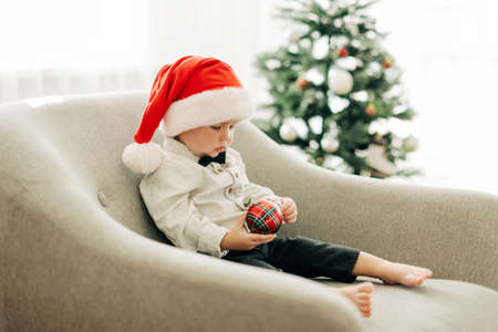 A little boy in a Santa Claus hat is sitting in an armchair with a Christmas tree toy in his hands next to the Christmas tree. Preparation for the holidays, festive decorの写真素材