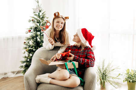 Two beautiful teenage girls in Christmas outfits open gifts at home. Christmas mood, Christmas gifts. The concept of a New Years holiday at homeの写真素材