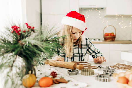 A girl in a red Christmas hat is making gingerbread dough in the kitchen. Christmas tradition, Christmas atmosphere, preparation for the holidayの写真素材