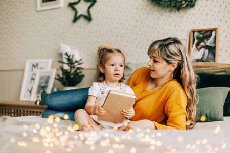 Mom and daughter are reading a book and smiling, sitting at home on the bed. New Year, Christmas, family, time togetherの写真素材
