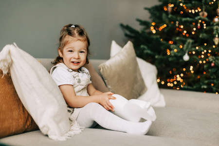 A pretty girl in a hat is sitting on the sofa near a Christmas tree, dressed up for the holiday, the child is smiling and rejoicing at the giftの写真素材