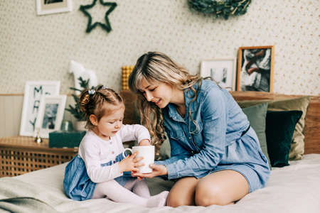 A smiling mother hides a gift for her daughter in a cup, congratulates her on Christmas, sitting on the bed on a winter holiday in a cozy houseの写真素材