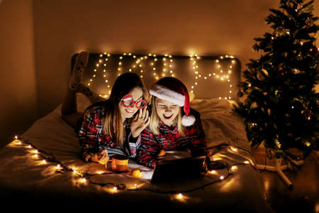 Two charming sisters lie on the bed with a laptop on Christmas or New Years eve and congratulate...