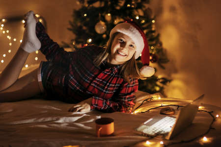 A teenage girl is lying on the bed next to a Christmas tree decorated for Christmas, using a laptop, there is a mug with a hot drink nearbyの写真素材