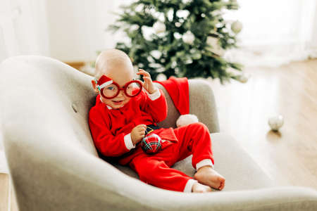 A charming toddler in decorative glasses, dressed as Santa Claus, is sitting in a chair next to a Christmas tree on Christmas Eveの写真素材