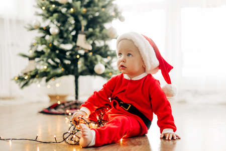A cheerful boy in a New Years costume sits on the floor near the Christmas tree and plays with a...