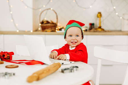 A little boy in a Christmas costume is sitting at a laptop in the kitchen. Congratulations online, Christmas wishes, virtual Christmasの写真素材
