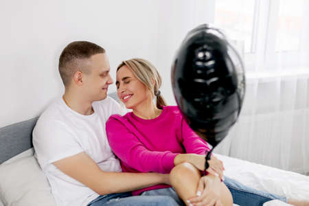 A young man hugs a girl sitting on a bed with a heart-shaped balloon during the celebration of Valentines Dayの写真素材