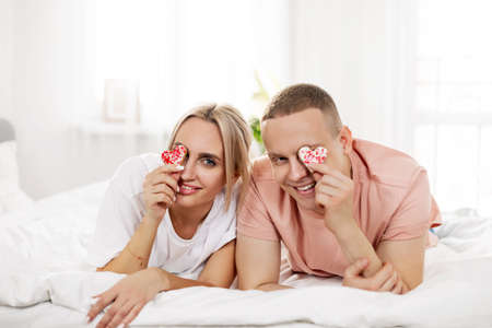 A young couple is lying on the bed, fooling around and covering their eyes with cookies while celebrating Valentines Day at home togetherの写真素材
