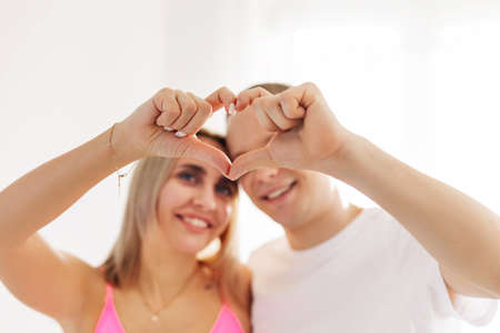 A couple stands close, hugging, making a heart shape with their hands and fingers, celebrating an...