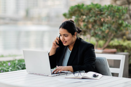 A young Indian woman is working on a laptop on the terrace in a cafe and talking on the phone. Online business, freelance.の写真素材