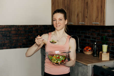 Portrait of an attractive woman holding a salad bowl and looking at the camera. Beautiful athletic girl in sportswear eating healthy salad after workout at homeの写真素材