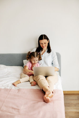 A young mother is working on a laptop or watching videos with her little daughter in the bedroom. Time together, work onlineの写真素材