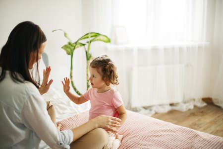 A young mother and a lovely daughter smile and clap their hands on the couch at home, playing...