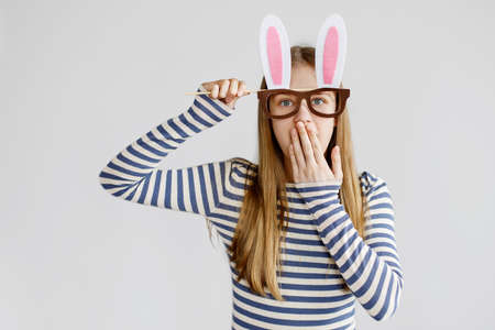 A teenage girl in a mask-glasses with rabbit ears in a striped sweater laughs and covers her mouth with her hand. Preparation for Easter. Easter bunny costume, Easter cardの写真素材