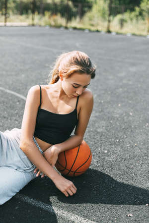 A basketball player is sitting on a sports field with a basketball and looks away. Sports, fitness, lifestyleの写真素材
