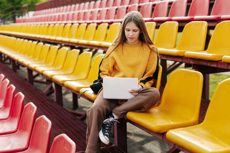 A young girl is doing her homework using a laptop in the stands of the stadium. The concept of training and educationの写真素材