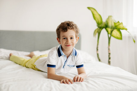 The cute boy is lying on his stomach on the bed on a white blanket, looking at the camera, smilingの写真素材