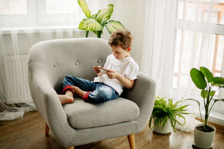 A cute boy is sitting in an armchair and playing a game on his phone or watching educational videos.の写真素材