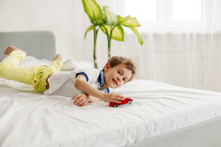 A charming boy is playing with a toy car on his bedの写真素材