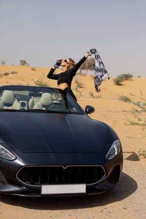 A glamorous girl in a stylish suit poses in a convertible car in the desert and waves a silk scarf....