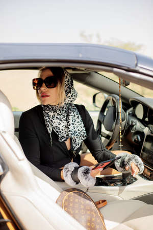A beautiful girl holding her smartphone in her hands is sitting in the car. A female driver reads a message and chats on a mobile phone.の写真素材