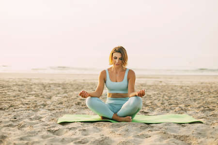 A happy young woman is sitting in a lotus position and doing yoga on the beach at sunset. The concept of a healthy active lifestyle.の写真素材