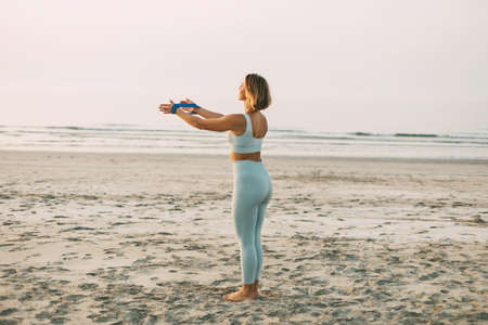 A slender girl performs hand exercises with a resistance band. A girl in sportswear trains on the beach.の写真素材