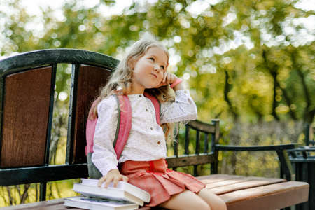 A cute thoughtful schoolgirl is sitting on a bench with books. Back to school, lesson schedule, elementary schoolの写真素材